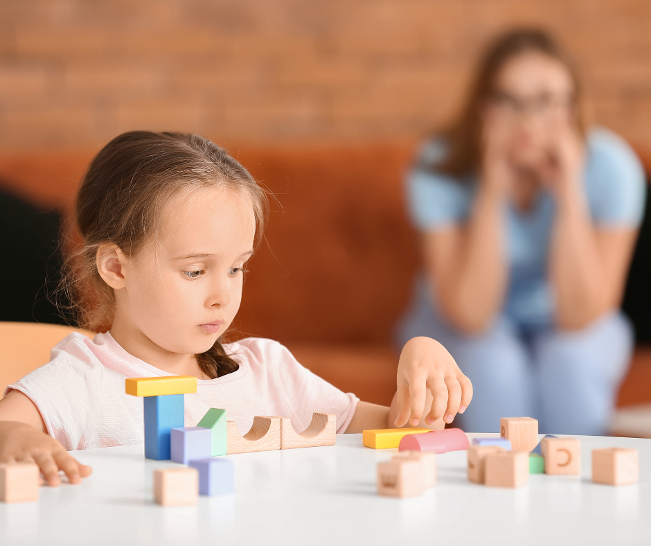 Photo of a 4 year old girl playing with blocks with her mom in the background holding her head with seeming concern.
