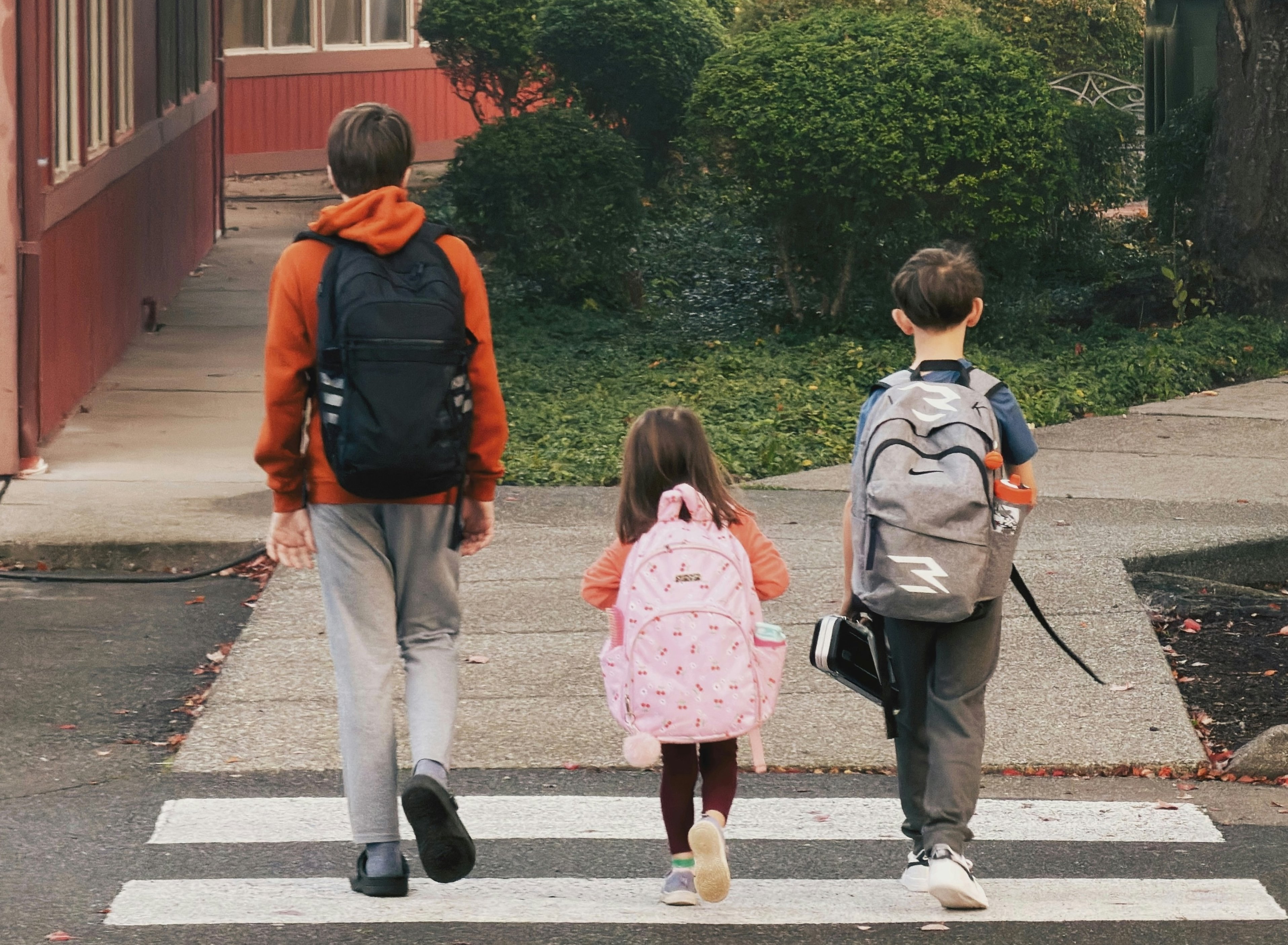 Three kids of varying ages wearing backpacks walking on a crosswalk in front of a school.