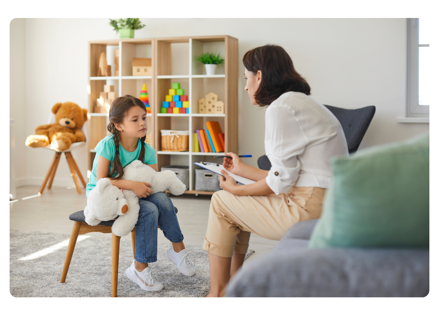 Brunette therapists chatting with 7 year old brunette girl in pig tails holding a stuffed bear in cheerful office.