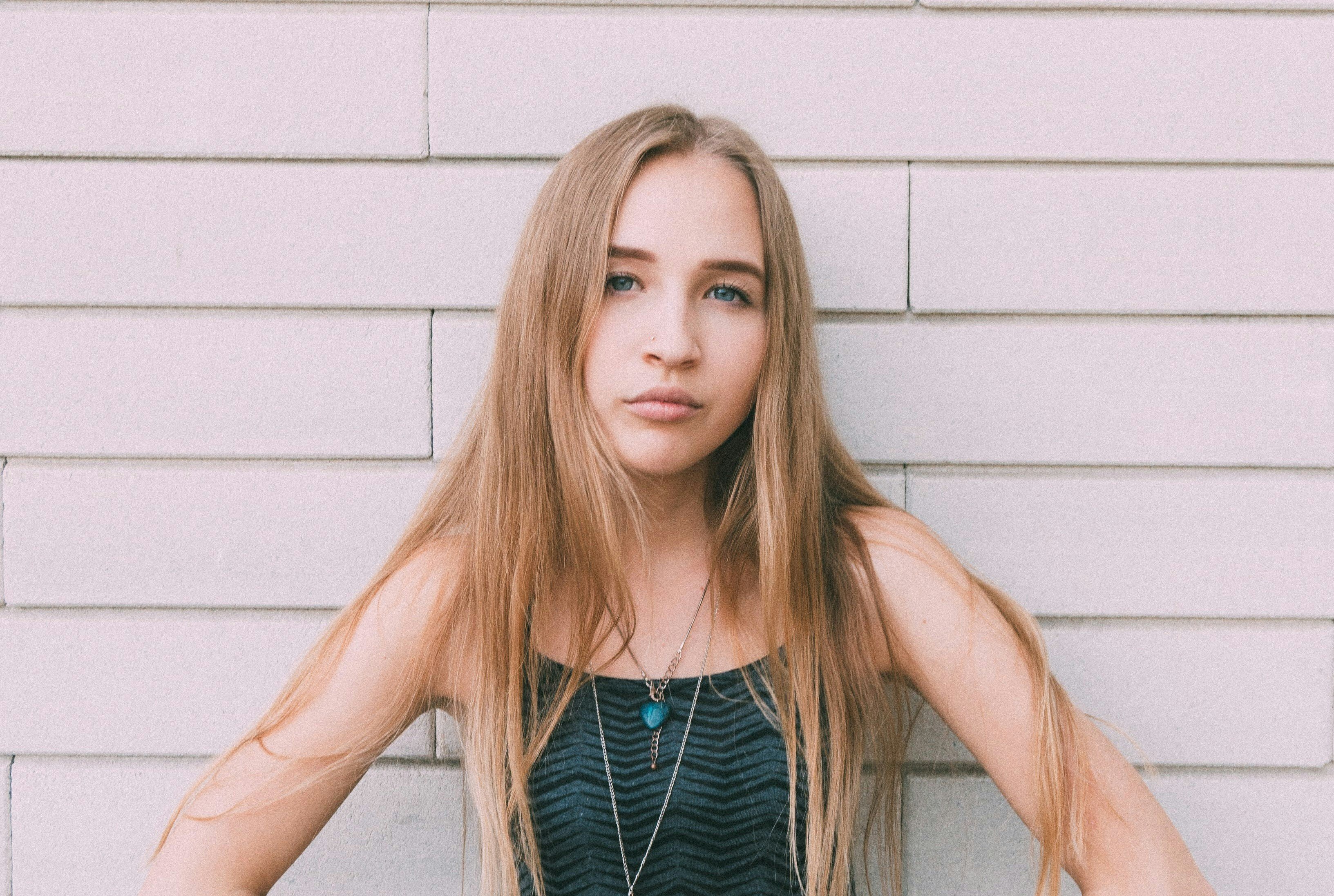 A teenage caucasian girl with a serious look on her face, standing in front of a wall of white bricks.