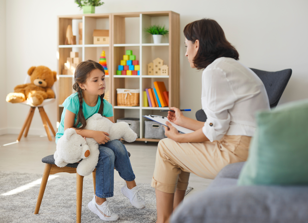 Female therapist chatting with a little girl, age 7, holding a stuffed toy bear in a cheerful well-lit office.