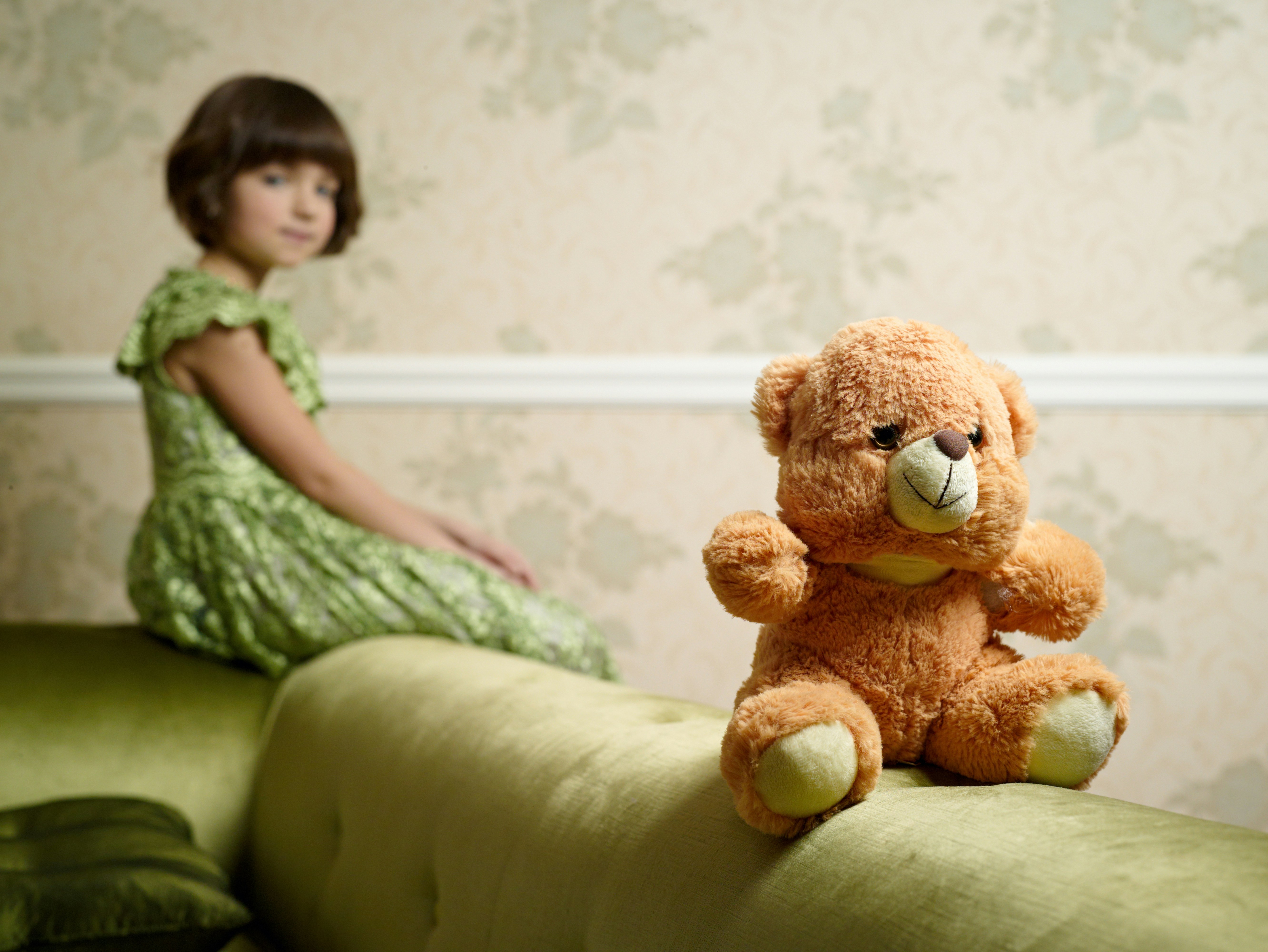 Six year old girl with a brunette bob wearing a green dress slightly out of focus in the background, her teddy bear is in focus in the foreground.