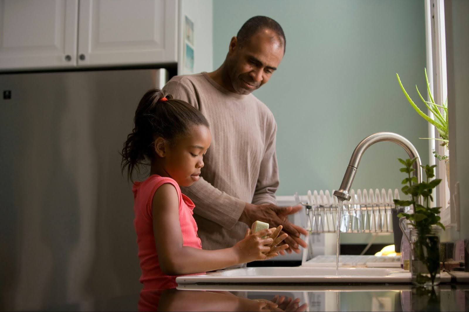 African American father and daughter standing at the sink doing dishes. Father is listening to or observing daughter.